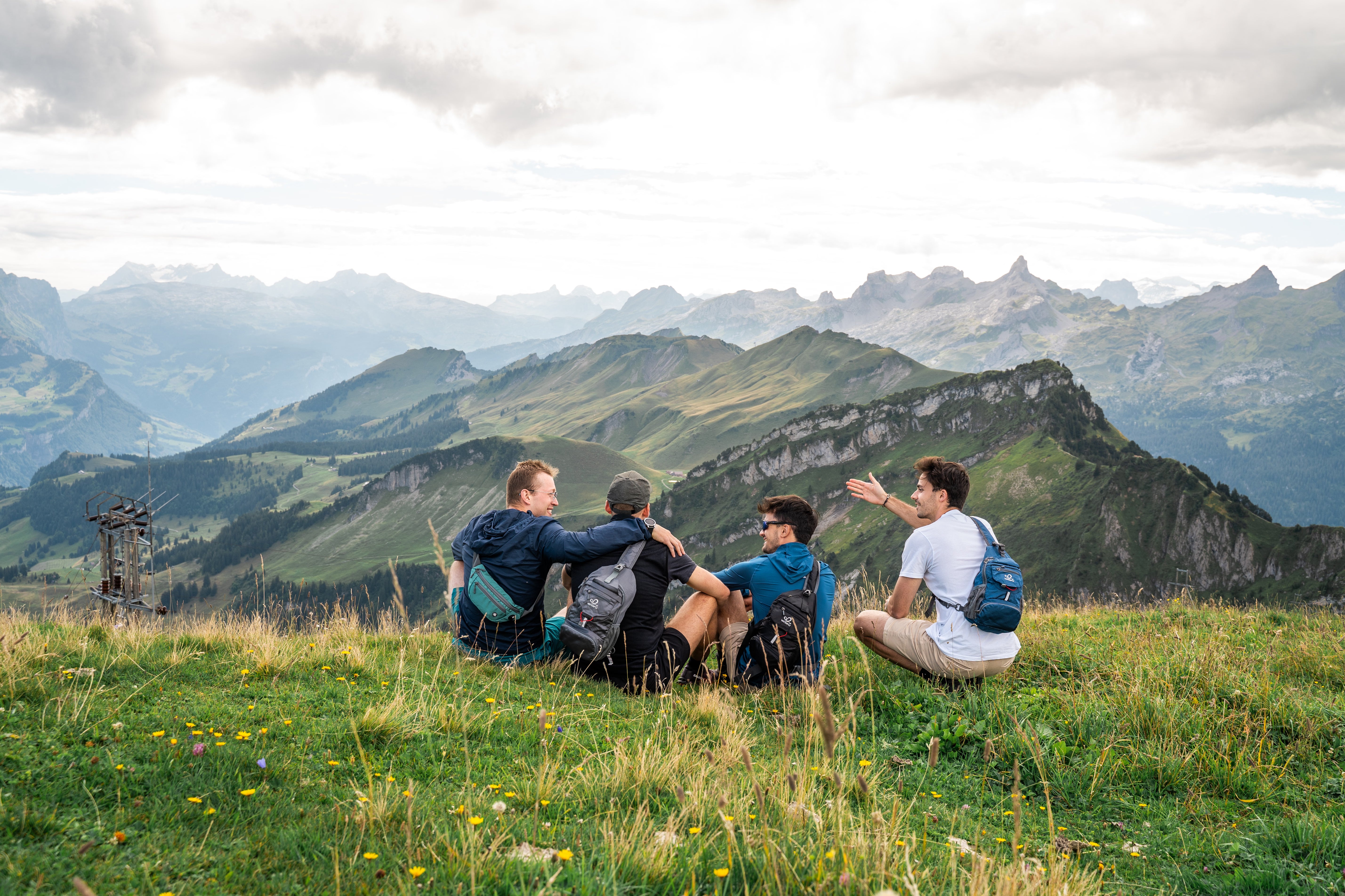 Four people with WATERFLY sling bags sitting on a grassy mountain viewpoint